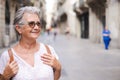 Happy senior woman walking in the city pedestrian street wearing sunglasses and backpack. Caucasian gray haired woman