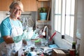 A happy senior woman while wash dishes. Gray hair and eyeglasses. wearing apron and gloves. Corner of kitchen. Grandmother at work