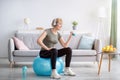 Happy senior woman working out with dumbbells on fitball and listeing to music at home, empty space