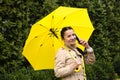Happy senior woman in yellow rain coat with yellow umbrella is talking on a smartphone and walking in park. Staying in
