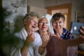 Happy senior women friends in bathrobes taking selfie indoors at home, selfcare concept.