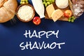 Happy Shavuot greeting card with dairy products, bread, fruits, wheat on blue background