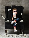 Happy and shoutting rich kid boy millionaire sits with a bundle of money dollars cash in big luxury armchair while bills falling Royalty Free Stock Photo