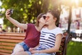 Happy sincere two girls make selfie on the wooden bench sitting in the park. Cheerful young girl kisses her best friend in right Royalty Free Stock Photo