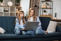 Happy sisters or friends two cute blond girls sitting together on comfortable couch, using laptop Royalty Free Stock Photo