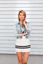 Happy slim young woman with a beautiful smile with blue eyes in a stylish white summer dress in a fashionable denim jacket posing