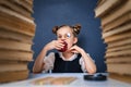 Happy smart girl in rounded glasses, eating a red apple while sitting between two piles of books and look at camera Royalty Free Stock Photo