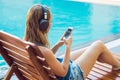 Happy smartphone woman relaxing near swimming pool listening with earbuds to streaming music. Beautiful girl using her Royalty Free Stock Photo