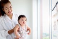 Happy smiling adorable six months baby girl standing next to the window in embrace of mother arms, mom holding her sweet little Royalty Free Stock Photo