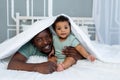 Happy smiling African american dad with baby son on the bed at home cuddling under the blanket, happy family, father`s day Royalty Free Stock Photo