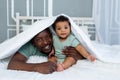 Happy smiling African american dad with baby son on the bed at home cuddling under the blanket, happy family, father`s day