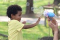 Happy smiling African boy with black curly hair playing with soap bubbles making from blowing bubble gun toy at green garden. Royalty Free Stock Photo