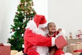 Happy smiling African child girl baby sitting on Santa Claus lap with decorative Christmas tree as background, cute kid open Royalty Free Stock Photo