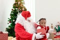 Happy smiling African child girl baby sitting on Santa Claus lap with decorative Christmas tree as background, cute kid open Royalty Free Stock Photo