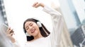 Happy Asian girl wearing headphones listening and moving to the music against city building background