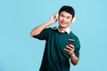 Happy smiling Asian man in green casual shirt wearing wireless headphones listening to music from smartphone isolated on light Royalty Free Stock Photo