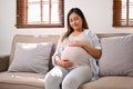 Happy Asian pregnant woman relaxes on the sofa in her living room, touching belly with love Royalty Free Stock Photo