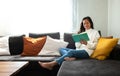 Happy, smiling Asian woman relaxing at home reading a book sitting on the sofa. Copy space. Royalty Free Stock Photo