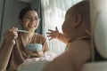 Happy Smiling Asian Young Mother enjoying feeding to her Little cute baby boy with a spoon while sitting on high chair at home. Royalty Free Stock Photo