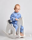 Happy smiling barefooted baby boy in blue fleece jumpsuit with stars plays rides white kids rocking horse toy