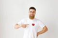 Happy smiling bearded male model wears casual white t shirt, round spectacles, indicates at red heart sewed on clothes Royalty Free Stock Photo