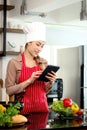 Happy smiling beautiful Asian woman wears apron and chef hat, holding and looking at digital tablet for searching tasty food meal Royalty Free Stock Photo