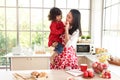 Happy smiling beautiful young Asian mom wearing cute red heart apron holding little daughter in lovely kitchen, mother feeding her
