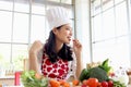 Happy smiling beautiful young Asian woman in red heart apron and chef hat holds and eats sliced carrot while sits behind kitchen Royalty Free Stock Photo