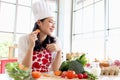 Happy smiling beautiful young Asian woman in red heart apron and chef hat holds and eats sliced carrot while sits behind kitchen Royalty Free Stock Photo