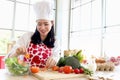 Happy smiling beautiful young Asian woman in red heart apron and chef hat holds sliced carrot for making salad while sits behind Royalty Free Stock Photo