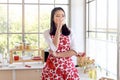 Happy smiling beautiful young Asian woman in red heart apron holds milk glass while stands behind kitchen counter. Girl relaxing Royalty Free Stock Photo