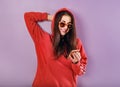 Happy smiling brunette woman of caucasian holding in hand and use mobile cell phone in fashion red hoodie and sunglasses on purple Royalty Free Stock Photo