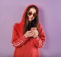 Happy smiling brunette woman of caucasian holding in hand and use mobile cell phone in fashion red hoodie and sunglasses on purple Royalty Free Stock Photo