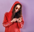 Happy smiling brunette woman of caucasian holding in hand and use mobile cell phone in fashion red hoodie and sunglasses on purple Royalty Free Stock Photo