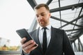 Happy smiling businessman wearing black suit and using modern smartphone near office at early morning, successful Royalty Free Stock Photo