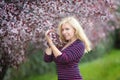 Happy smiling Caucasian blond woman with long hair in purple fedora hat near blossoming plum cherry tree, enjoys the blossom Royalty Free Stock Photo