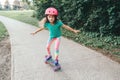 Happy smiling Caucasian girl in pink helmet riding skateboard on road in park on summer day. Seasonal outdoors children activity Royalty Free Stock Photo