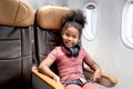 Happy smiling curly hair African girl child passenger with headphones sitting in seat next to window inside airplane, cheerful kid Royalty Free Stock Photo