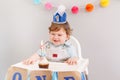 Happy smiling cute Caucasian baby boy in blue crown celebrating his first birthday at home. Child kid toddler sitting in high Royalty Free Stock Photo