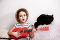 Happy smiling cute little girl in hat singing and playing small guitar ukulele at home. Funny black cat at background listening Royalty Free Stock Photo