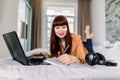 Happy smiling dark haired woman lefthander using laptop and making notes in her diary notepad, while lying on bed in the Royalty Free Stock Photo