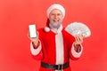 Happy smiling elderly man with gray beard wearing santa claus costume holding fan of money and showing smartphone with white blank Royalty Free Stock Photo