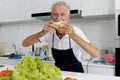 Happy smiling elderly man wearing apron, standing at kitchen counter. Senior man holding bread sandwich and ready to eat it, Royalty Free Stock Photo