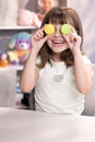 Happy smiling face of little girl covering her eyes with macaroons. Teen girl plays with dessert macarons, holding the