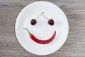 Happy smiling face made of cherry and chili pepper on white plate. Top view, wooden table overhead high angle above view photo. We