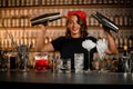 Happy smiling female bartender holding two cocktail shakers in front of various glasses