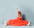 Happy girl with colorful dreadlocks in coral dress, sneakers, sunglasses sits on floor legs wide apart holding finger up Royalty Free Stock Photo