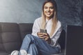 Happy smiling girl sitting on sofa, holding smartphone in hand. Young woman installing messenger app to communicate Royalty Free Stock Photo