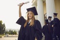 Happy smiling graduate girl dressed in black gown raises up her hand holding a diploma. Royalty Free Stock Photo