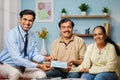 Happy smiling indian banker giving cheque to senior couple by looking at camera at home - concept of loan approval